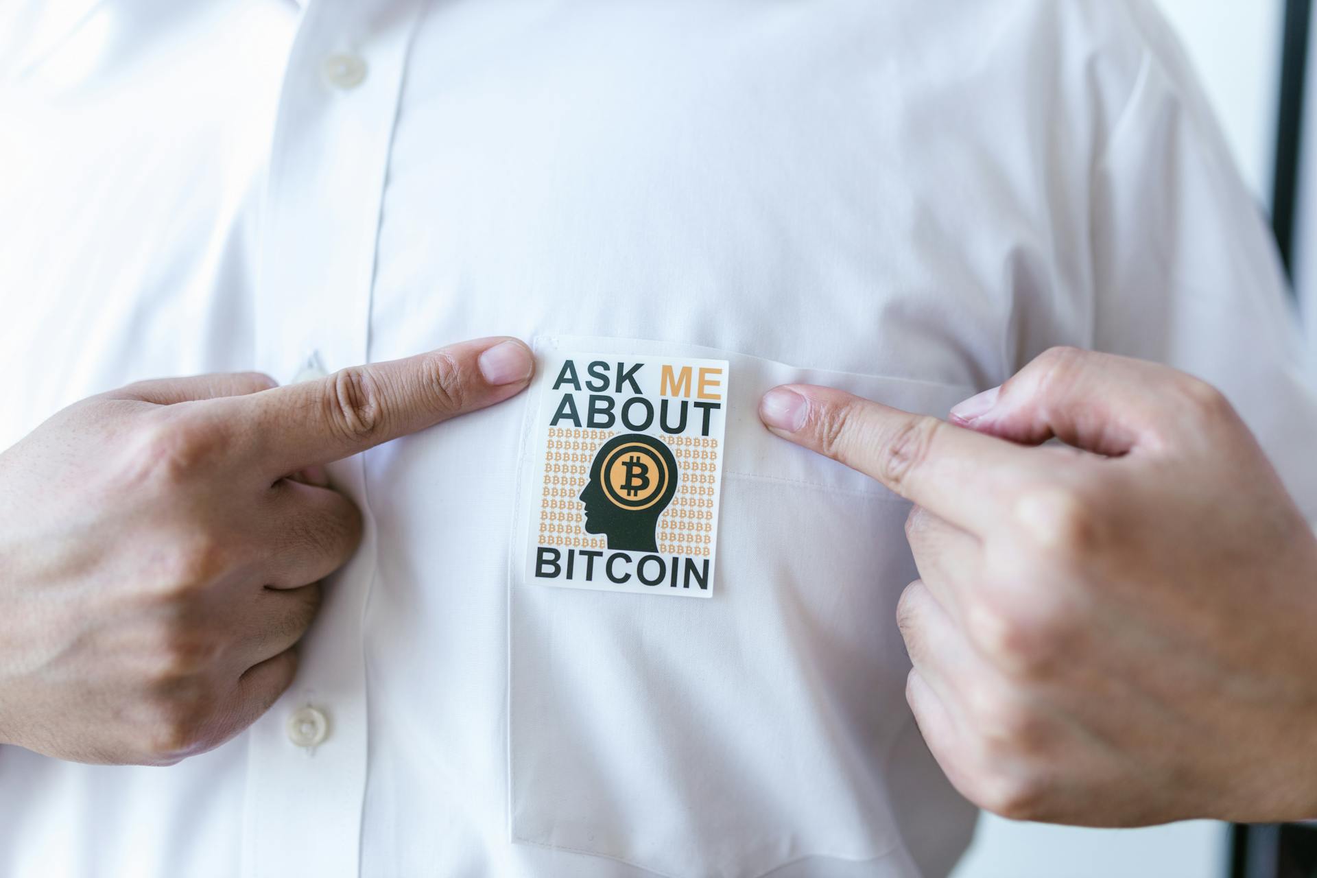 man with white shirt, his fingers pointing at a label tat says "Ask me about Bitcoin"